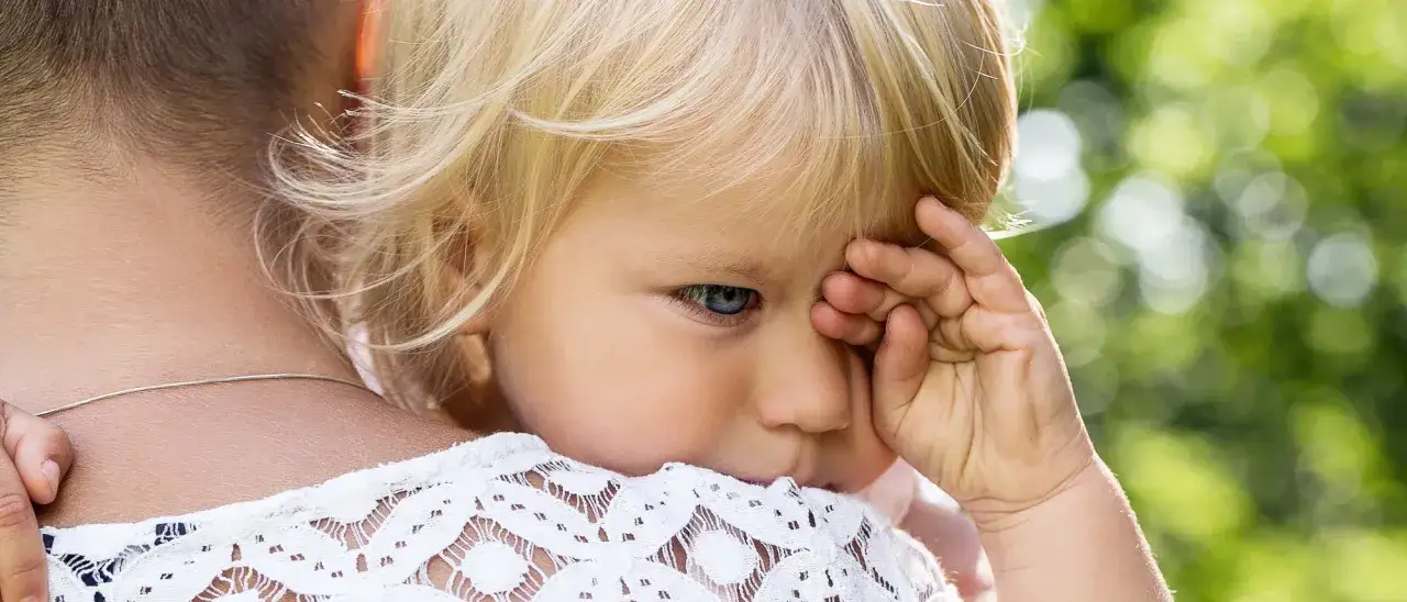Ein 2-j&auml;hriges Kind mit blonden Haaren und blauen Augen schaut traurig, die Hand vor dem Gesicht. Mama, bitte geh nicht weg!