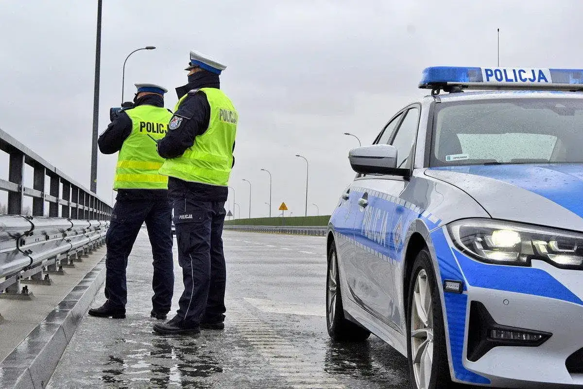 Dwóch policjantów w kamizelkach odblaskowych stoi przy radiowozie. Jeden z nich trzyma miernik prędkości, gotowi do akcji przeciwko niezatrzymaniu się do kontroli.