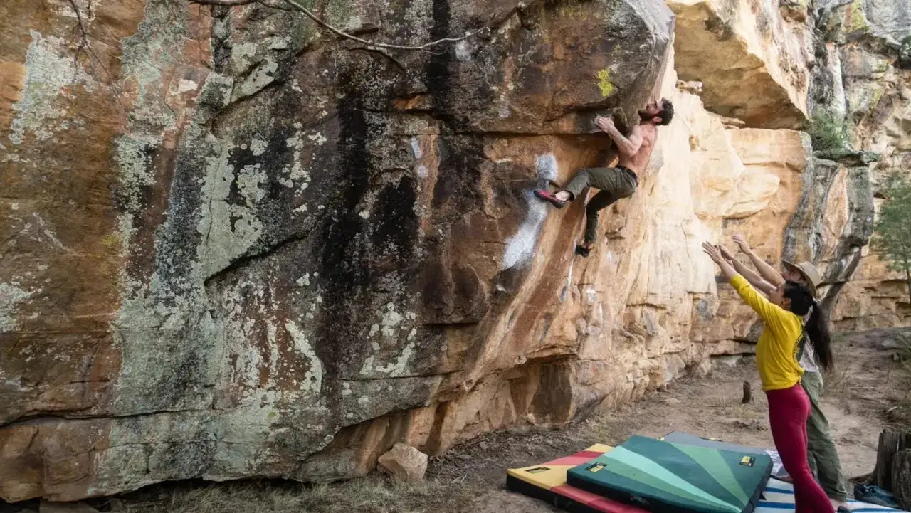 Mężczyzna wspina się po skale, podczas gdy jego przyjaciele asekurują go na dole. To ekscytująca sesja boulderowa.