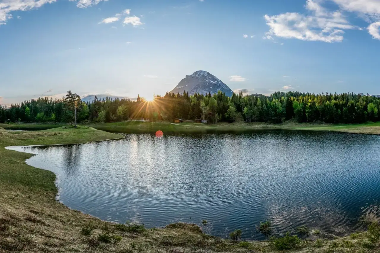 Sonnenaufgang &uuml;ber einem Bergsee in den Karnischen Alpen, umgeben von gr&uuml;nen W&auml;ldern und Wiesen.