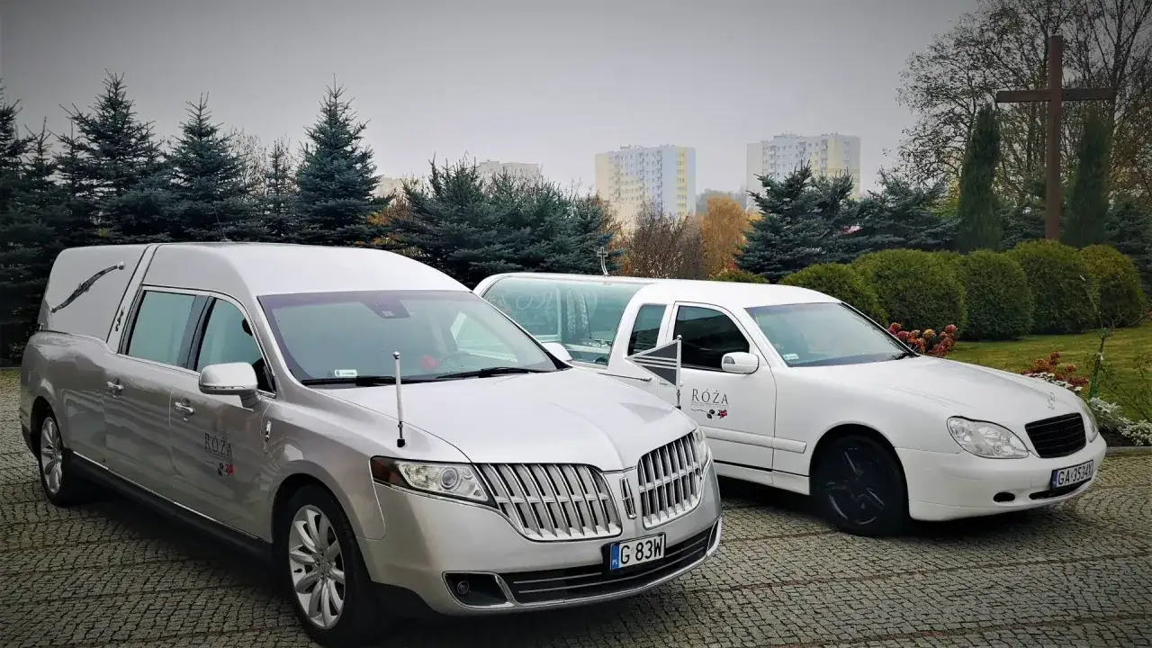 Dwa karawany, srebrny i biały, gotowe do transportu zwłok powyżej 60 km. Tło stanowi cmentarz z drzewami i budynkami.