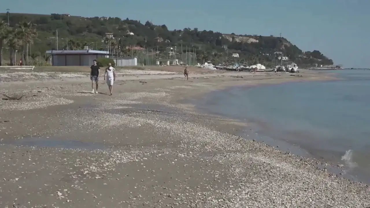 Passeggiata sulla spiaggia di Marina di Campofilone, con due uomini che camminano e barche all'orizzonte.