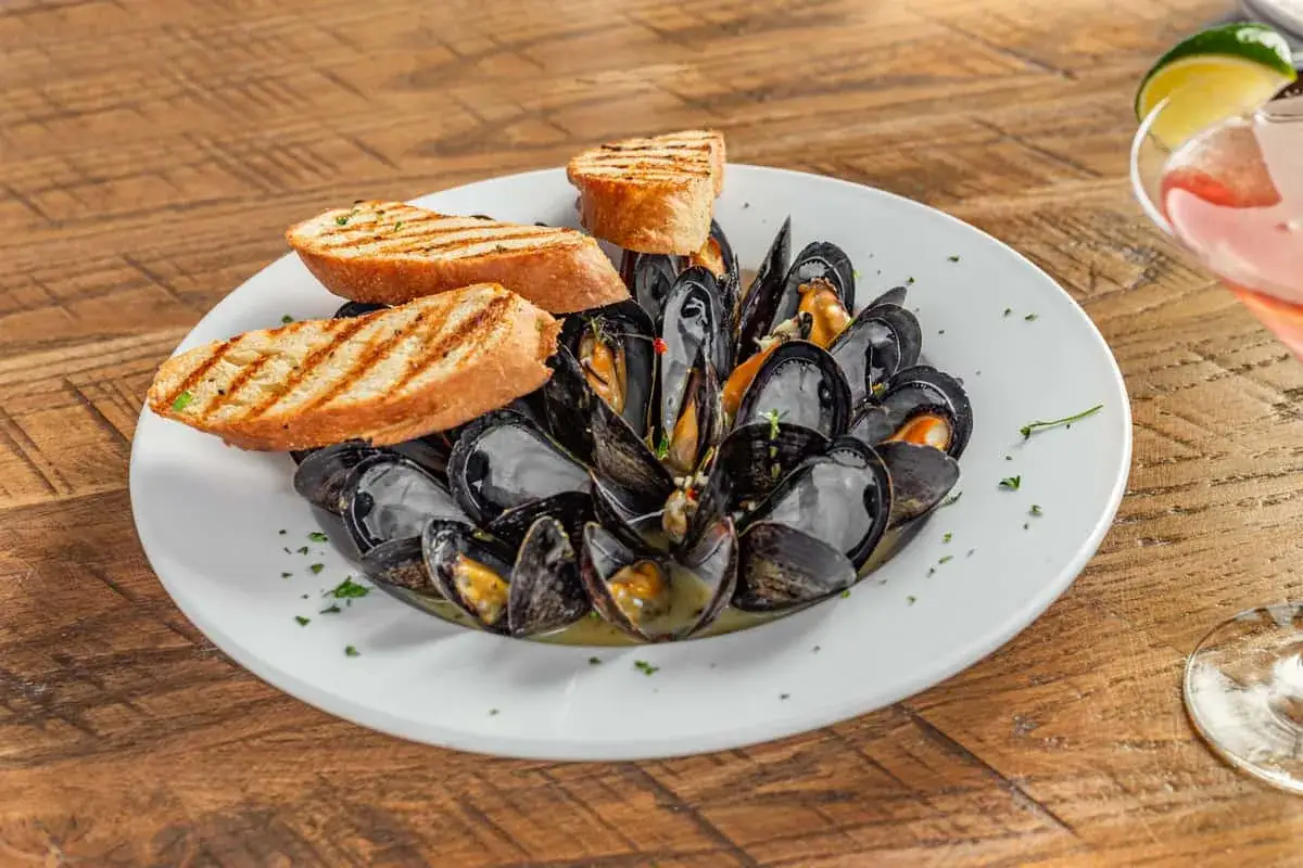 A steaming plate of moules marinière, brimming with dark mussels in a savory broth, is garnished with grilled bread slices.