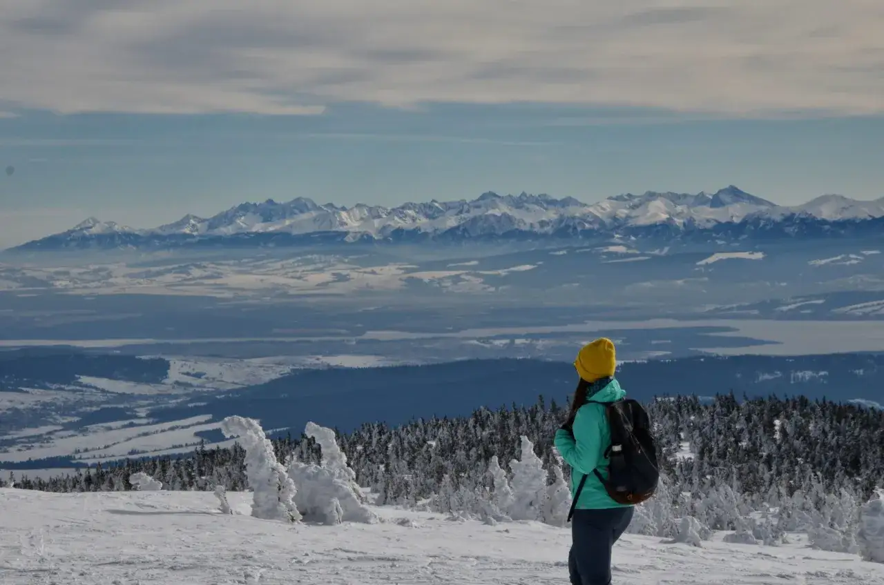Widok z **pilsko szczyt** na ośnieżone góry. Kobieta w żółtej czapce podziwia panoramę.