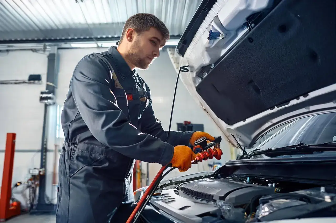 Mécanicien avec des gants orange manipulant un appareil pour la recharge clim voiture. Le capot est ouvert.