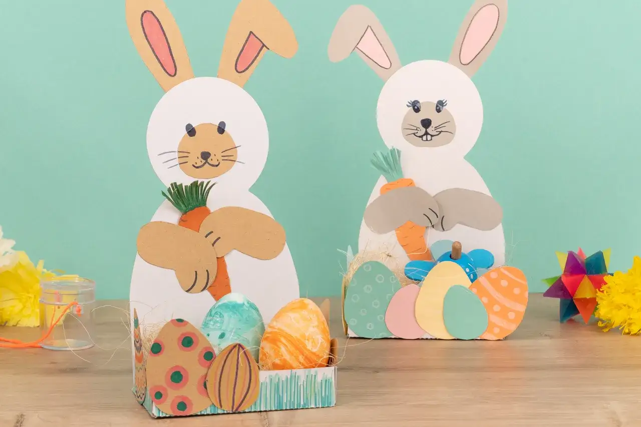 Zwei s&uuml;&szlig;e Osterhasen zum osterhase basteln halten Karotten. Bunte Ostereier liegen in K&ouml;rbchen.