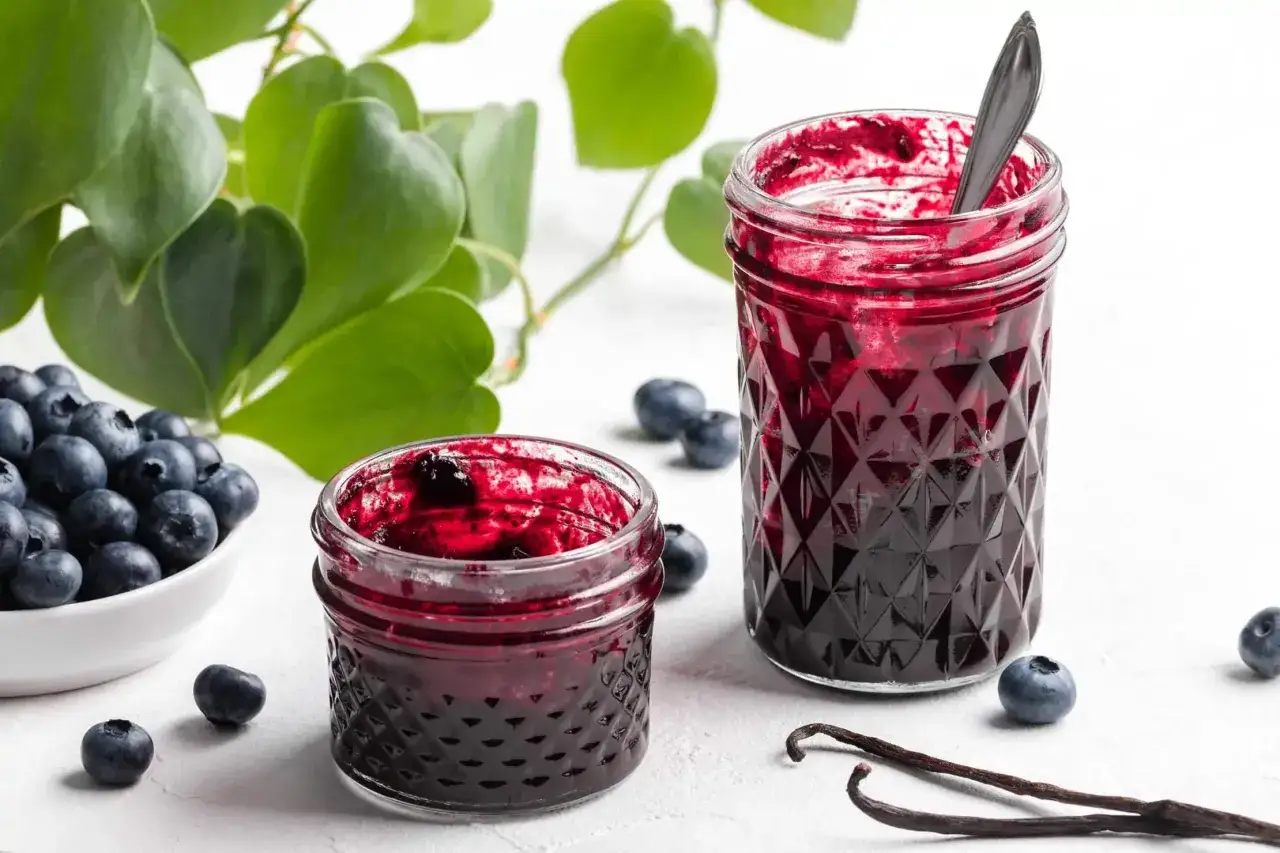 Frische Heidelbeeren und selbstgemachte Heidelbeermarmelade in Gläsern, daneben Vanilleschoten und grüne Blätter.