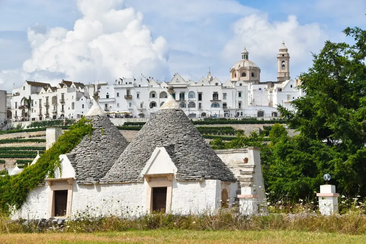 Charming trulli houses with conical roofs in the foreground, with a white hillside town and domed church in the background. A perfect place to base yourself in Puglia.