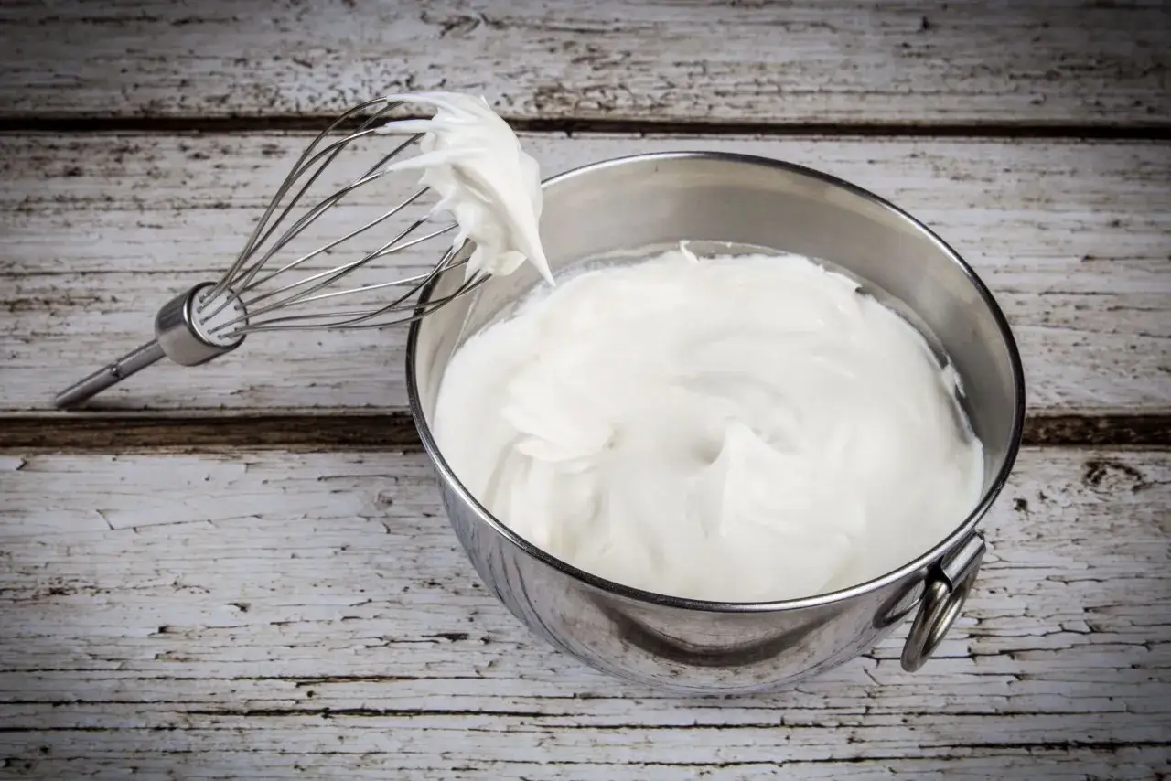 Ecco come fare la panna montata in casa: una frusta con panna montata soffice sopra una ciotola di panna montata, su un tavolo di legno.