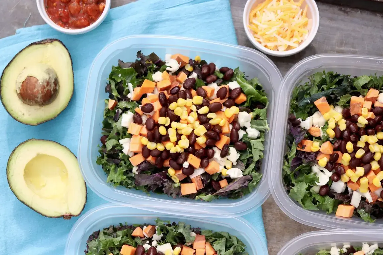 Vorbereitete Mahlzeiten f&uuml;r clean eating rezepte: Salate mit Gr&uuml;nkohl, S&uuml;&szlig;kartoffeln, schwarzen Bohnen und Mais in Beh&auml;ltern. Daneben Avocado und Salsa.