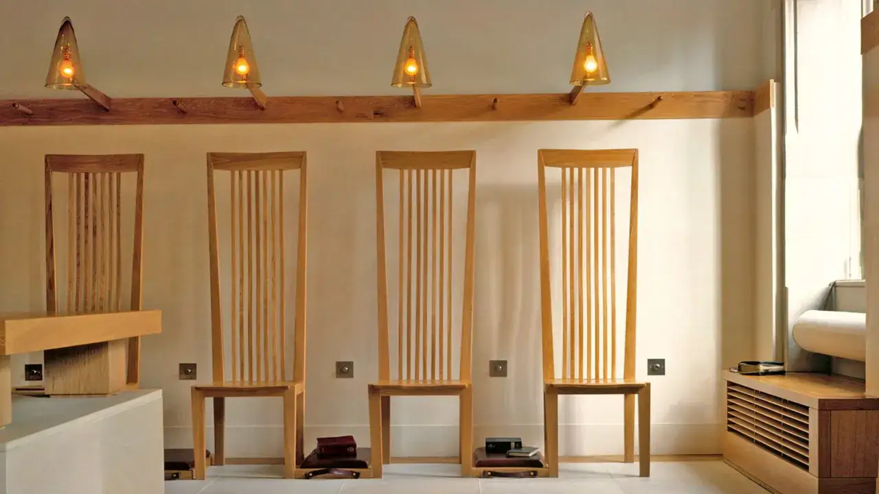 Four tall wooden chairs await guests, embodying Benedictine hospitality. Warm light from cone-shaped lamps illuminates the serene space.