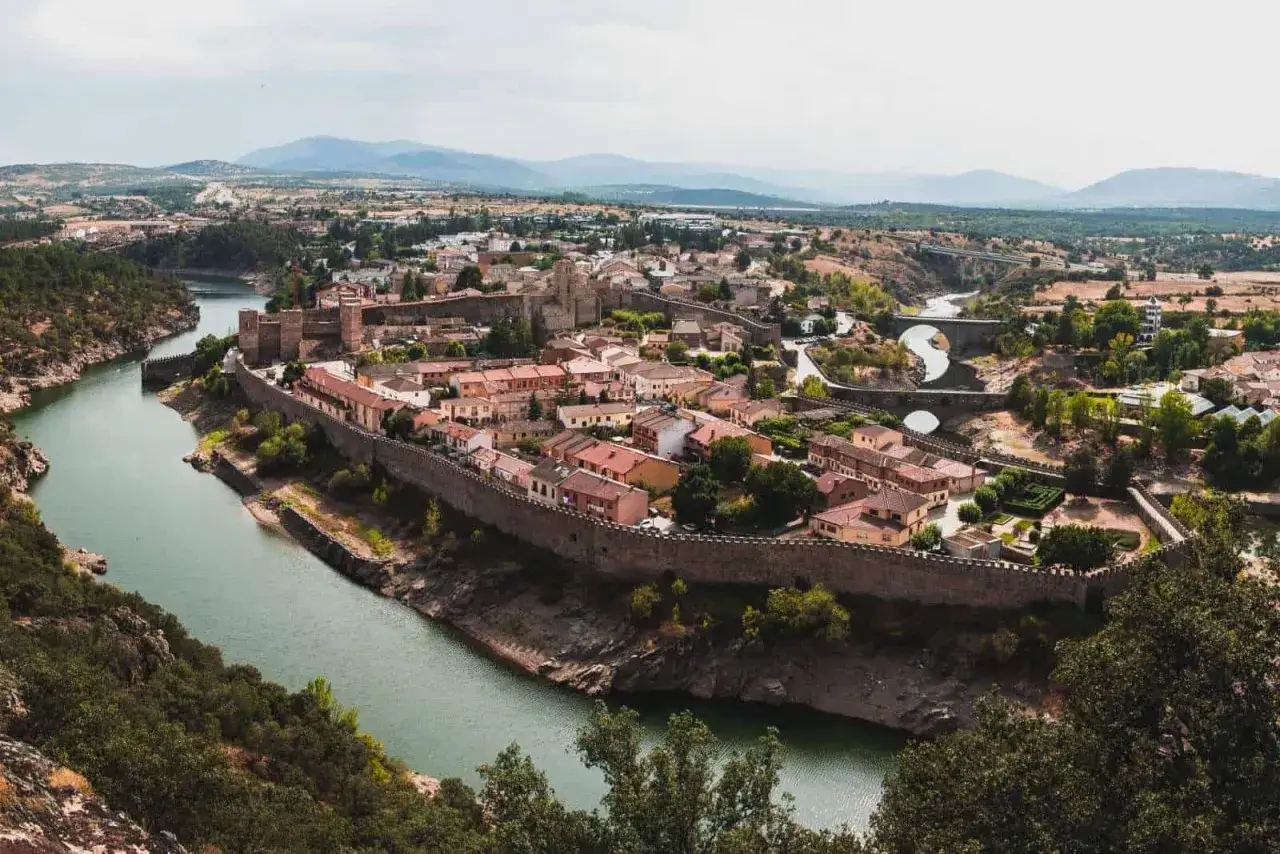 Vista panor&aacute;mica de Buitrago de Lozoya, un pueblo amurallado junto a un r&iacute;o, con un puente y monta&ntilde;as al fondo.