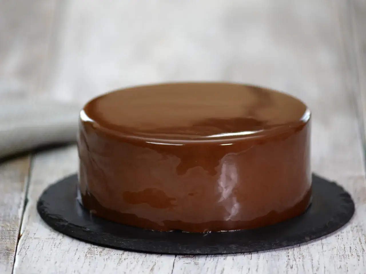 Gâteau rond avec un glaçage chocolat miroir brillant, posé sur une assiette ardoise.
