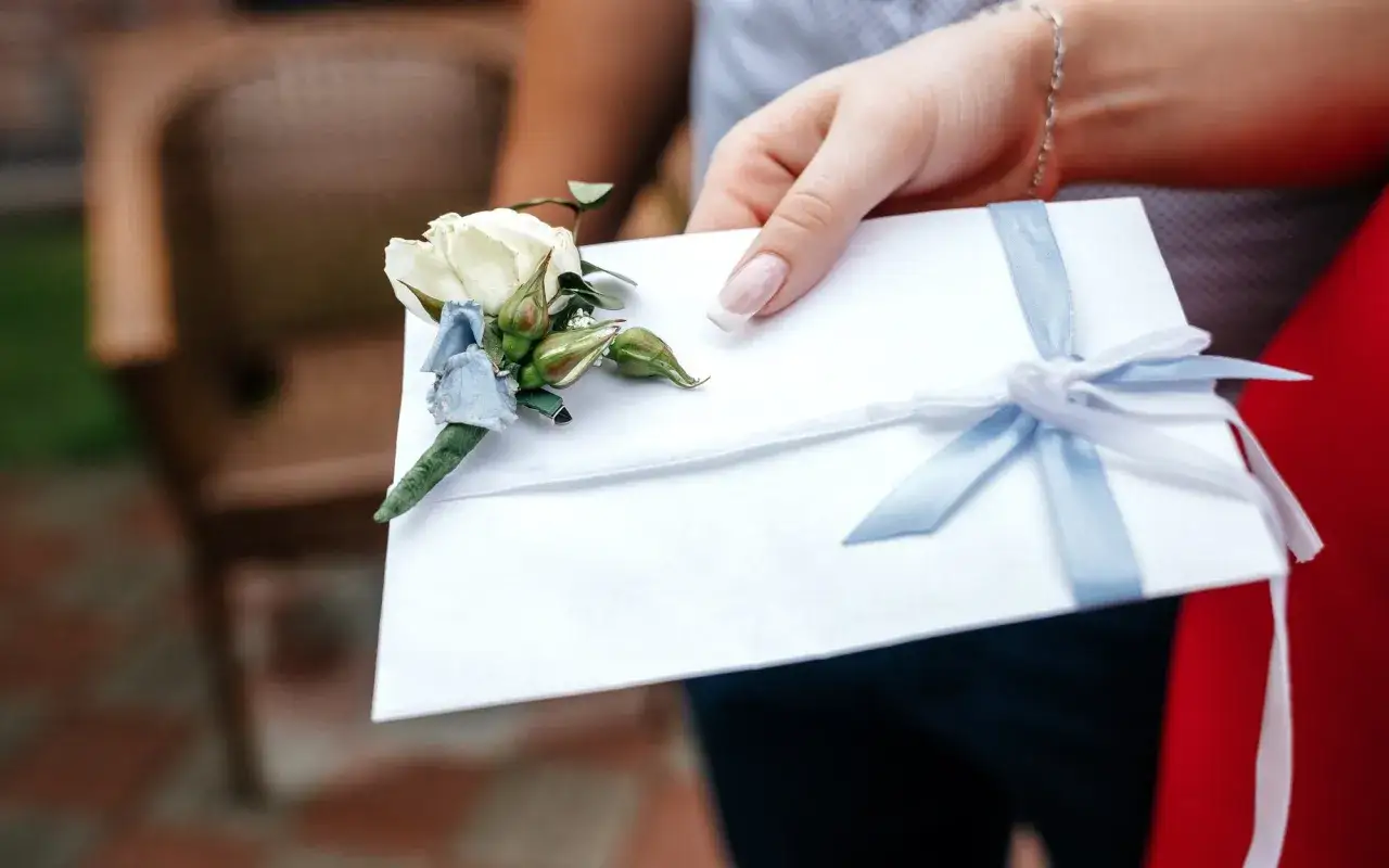 Dłoń z paznokciami w stylu francuskiego manicure trzyma białą kopertę ozdobioną bukiecikiem i błękitną wstążką. To prezent ślubny, który trafi na konto.