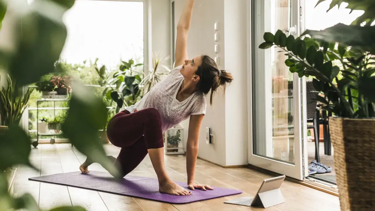 Kvinna utf&ouml;r yogaposition framf&ouml;r en surfplatta, som om hon f&ouml;ljer en yoga dvd.