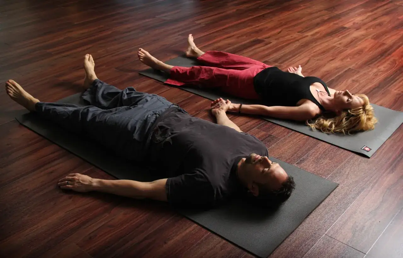 Pareja en Savasana, una de las posturas para hacer con tu pareja, relajándose en esterillas de yoga sobre suelo de madera.