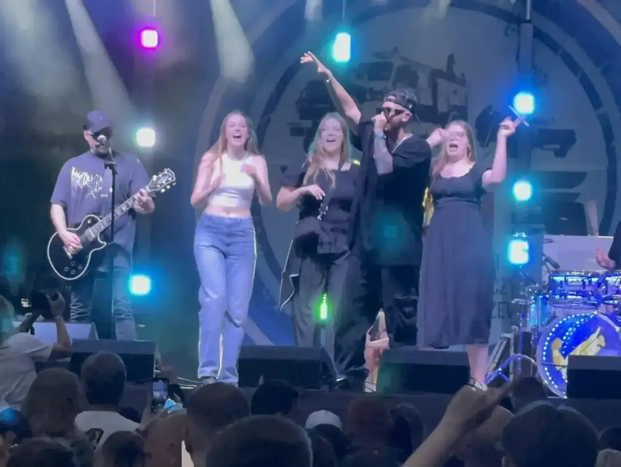 Zespół na scenie wykonuje utwory. Wokalista w czapce i okularach, gitarzysta i trzy młode kobiety śpiewają i tańczą.