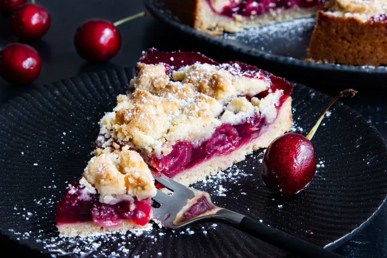 Ein saftiges St&uuml;ck von Omas Kirschkuchen mit Streuseln, best&auml;ubt mit Puderzucker. Daneben liegen frische Kirschen.