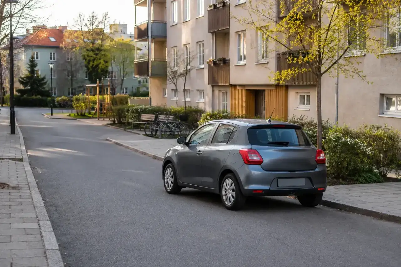 Grå Suzuki Swift står parkerad vid en trottoar framför ett bostadshus. Kanske ett problem med parkeringen?