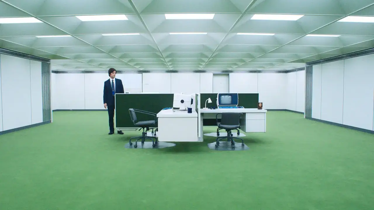 Uomo in abito scuro in un ufficio vuoto con moquette verde. La scissione trama del soffitto crea un senso di disorientamento.