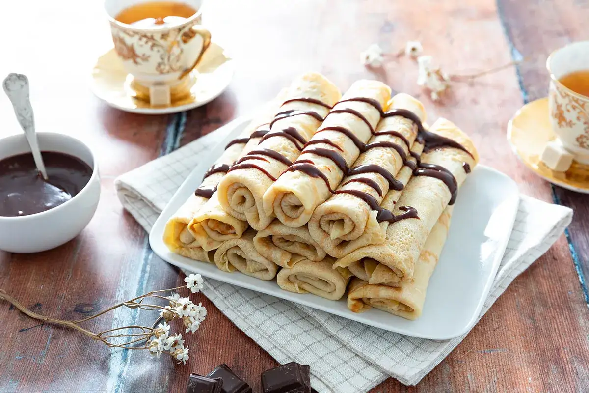 Pile de crêpes moelleuses arrosées de chocolat, accompagnées d'une tasse de thé et d'une coupelle de sauce chocolat. Une invitation à la gourmandise.