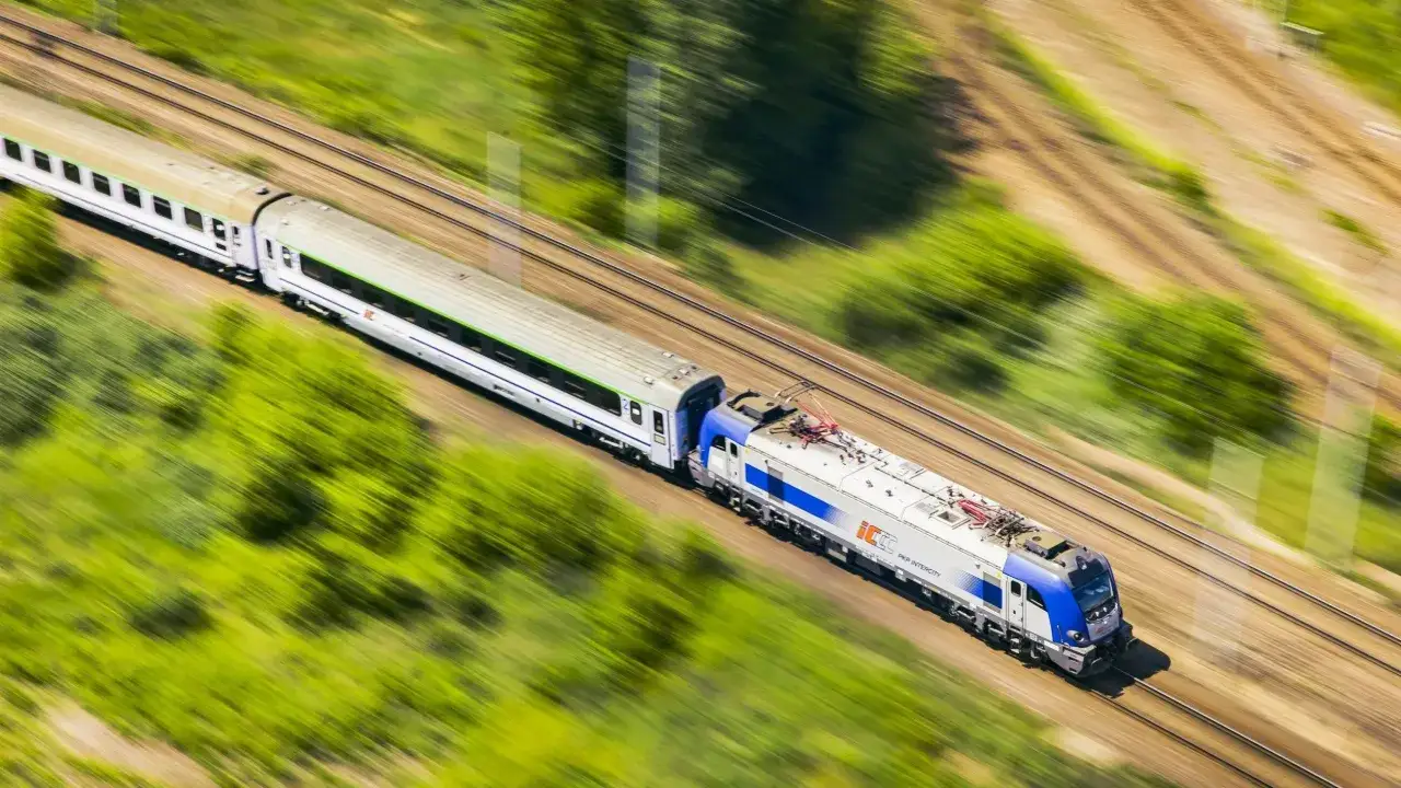 Pędzący pociąg PKP Intercity, model EU07, z białymi wagonami i zielonym pasem, mija zielone drzewa.