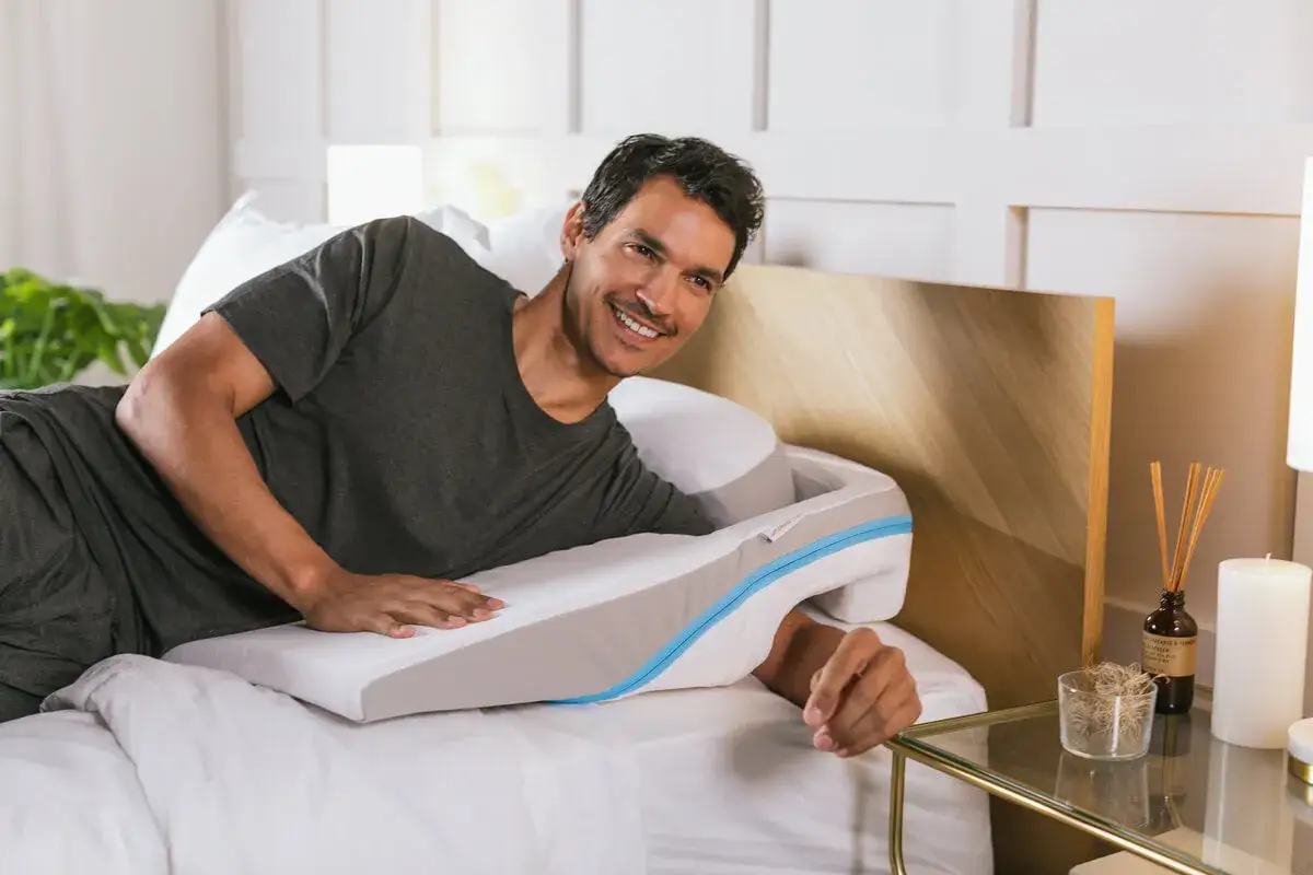 Man smiles in bed, resting on a supportive pillow designed for neck pain relief. These almohadas para dolor de cuello offer comfort and alignment.