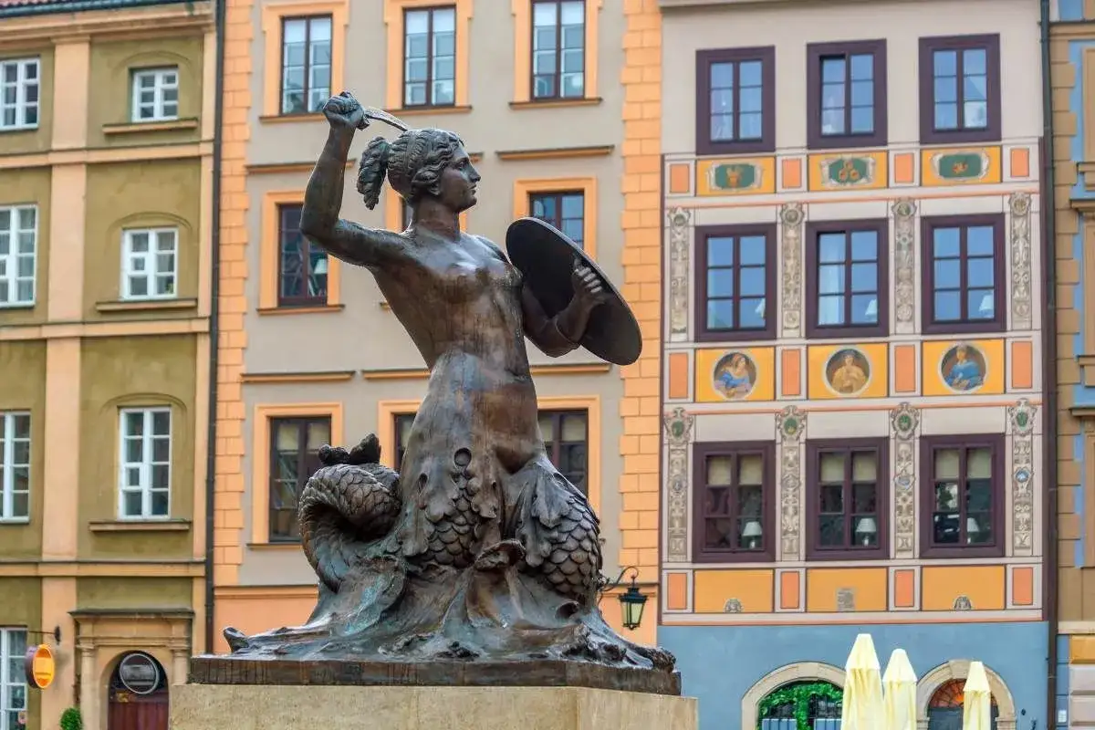 Pomnik Syrenki Warszawskiej, bohaterki legendy o Warsie i Sawie, z mieczem i tarczą, na tle kolorowych kamienic.