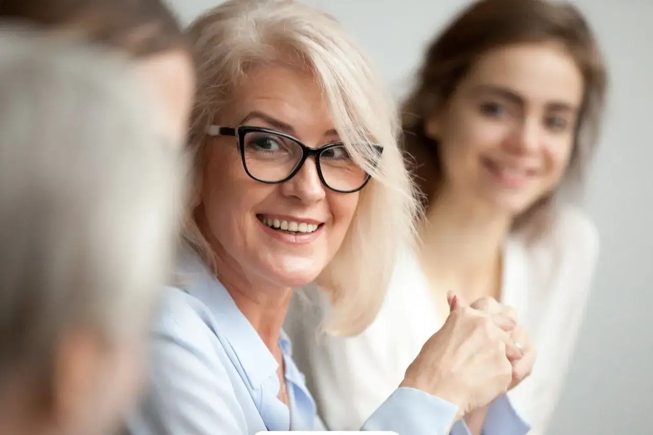Donna sorridente con occhiali partecipa a un mentoring aziendale, condividendo la sua esperienza con un collega più giovane.