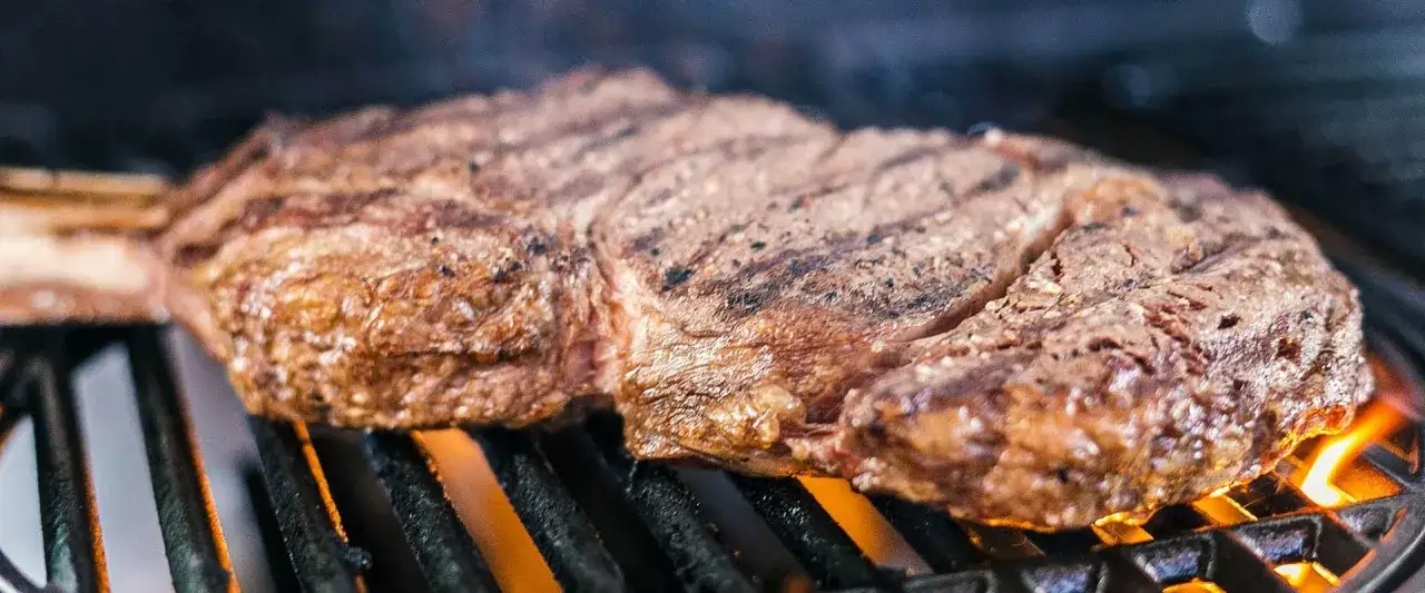 Saftiges Steak wird &uuml;ber gl&uuml;hender Holzkohle gegrillt, die Flammen z&uuml;ngeln unter dem Rost hervor.