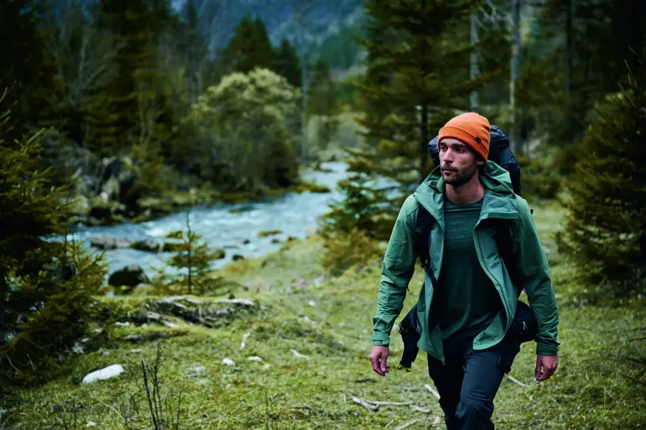 Escursionista con zaino indossa un impermeabile montagna, cammina lungo un torrente in un bosco.