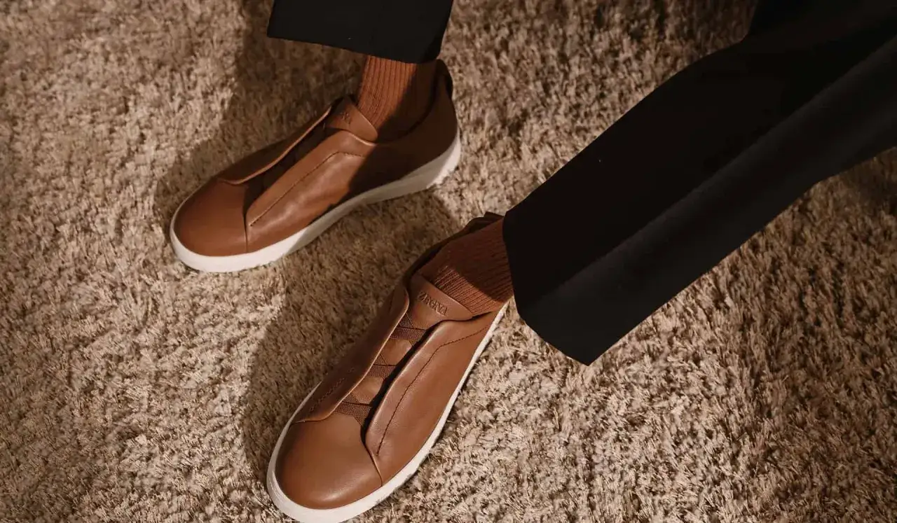 Elegante braune Leder-Sneaker mit weißen Sohlen, getragen mit braunen Socken und schwarzen Hosen. Diese 1000 € teuersten Schuhe der Welt strahlen Luxus aus.