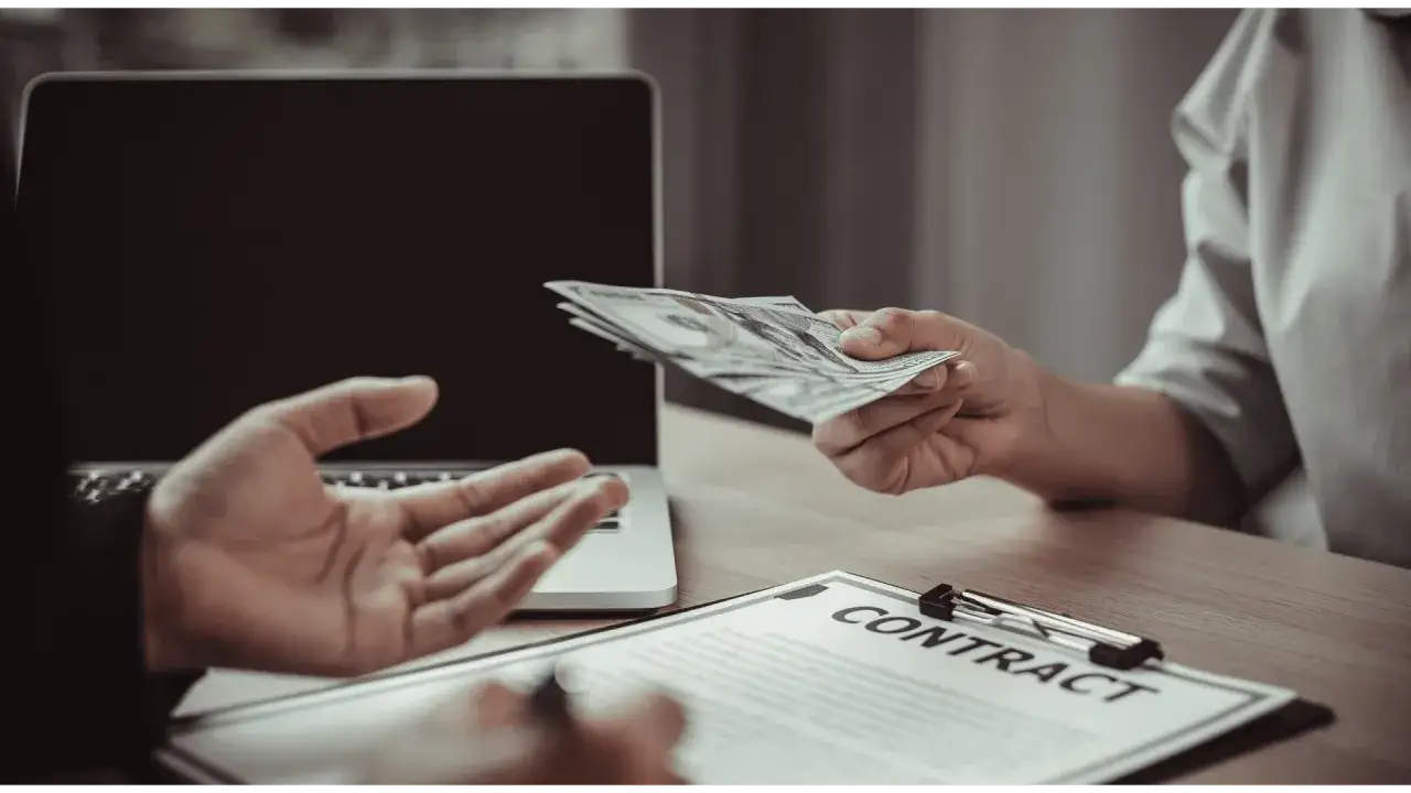 Dwie osoby przy biurku z laptopem i umową. Jedna podaje drugiej plik banknotów. Szybki kredyt na tak przez telefon.