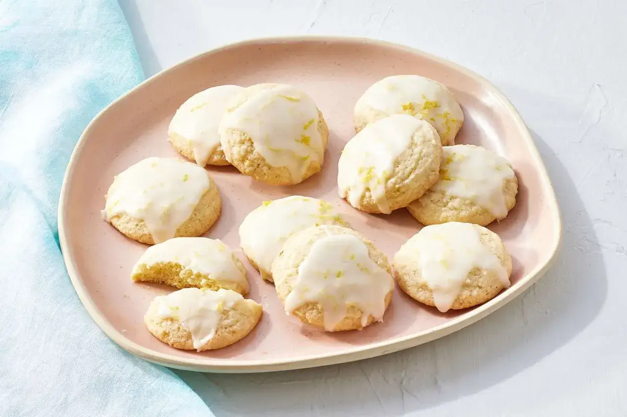 Zitronen Cookies mit Zuckerguss und Zitronenschale auf einem rosa Teller. Ein Keks ist zerbrochen und zeigt die zarte Textur.