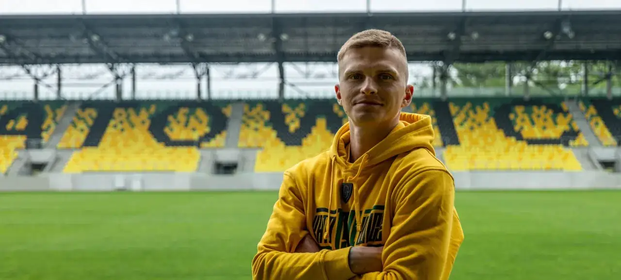 Piłkarz GKS Katowice w żółtej bluzie z kapturem na tle stadionu. Nowe transfery wzmocnią drużynę.