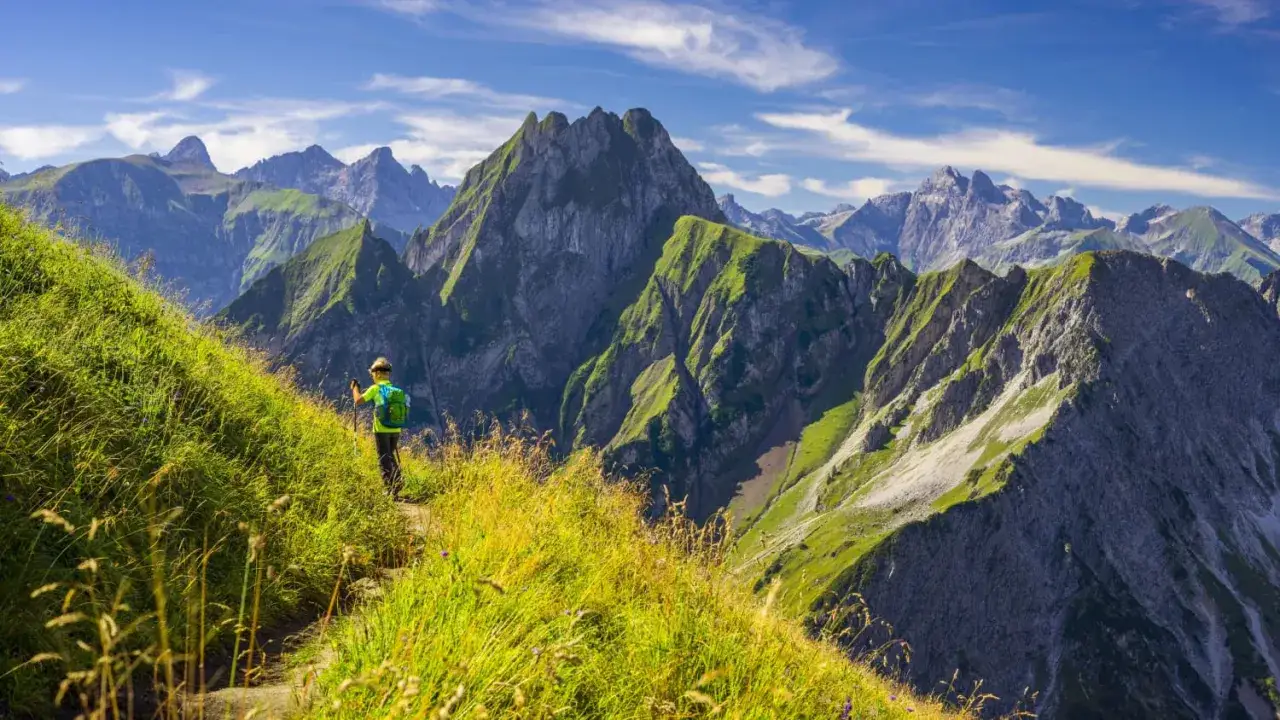 Ein Wanderer genießt die atemberaubende Aussicht auf die Berge beim Wandern in Deutschland. Grüne Almwiesen und schroffe Gipfel unter blauem Himmel.