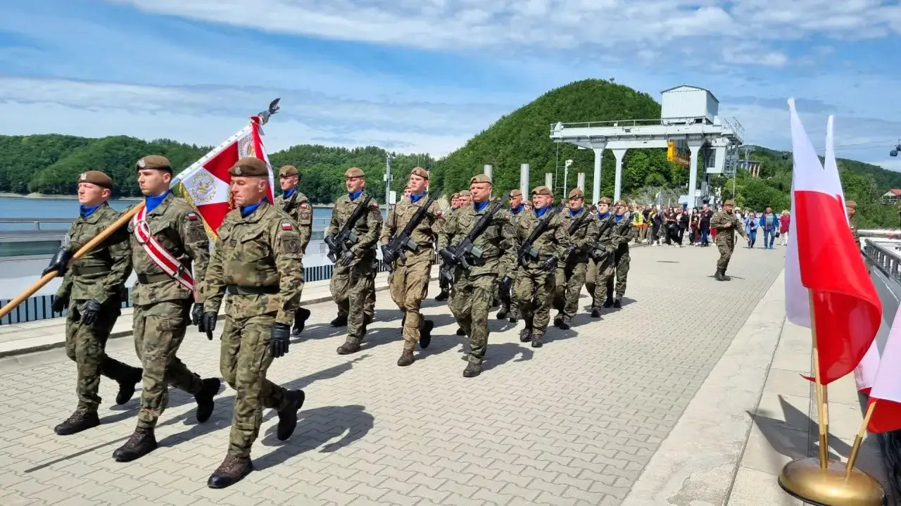 Żołnierze w pełnym rynsztunku maszerują z flagą, składając **przysięgę** w malowniczej scenerii nad jeziorem.