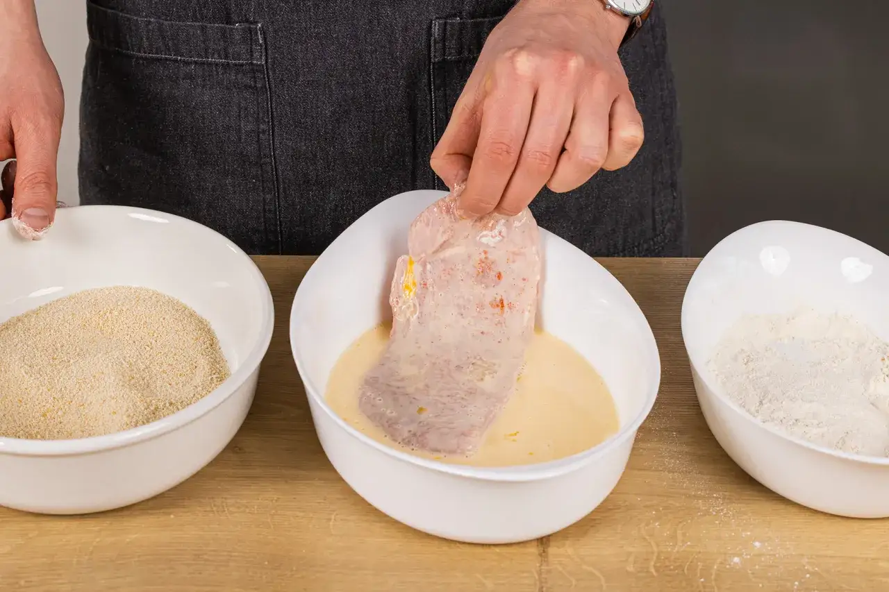 Schneiden wird in Ei getaucht, um es f&uuml;r das **Schnitzel panieren** vorzubereiten. Daneben stehen Sch&uuml;sseln mit Mehl und Paniermehl.