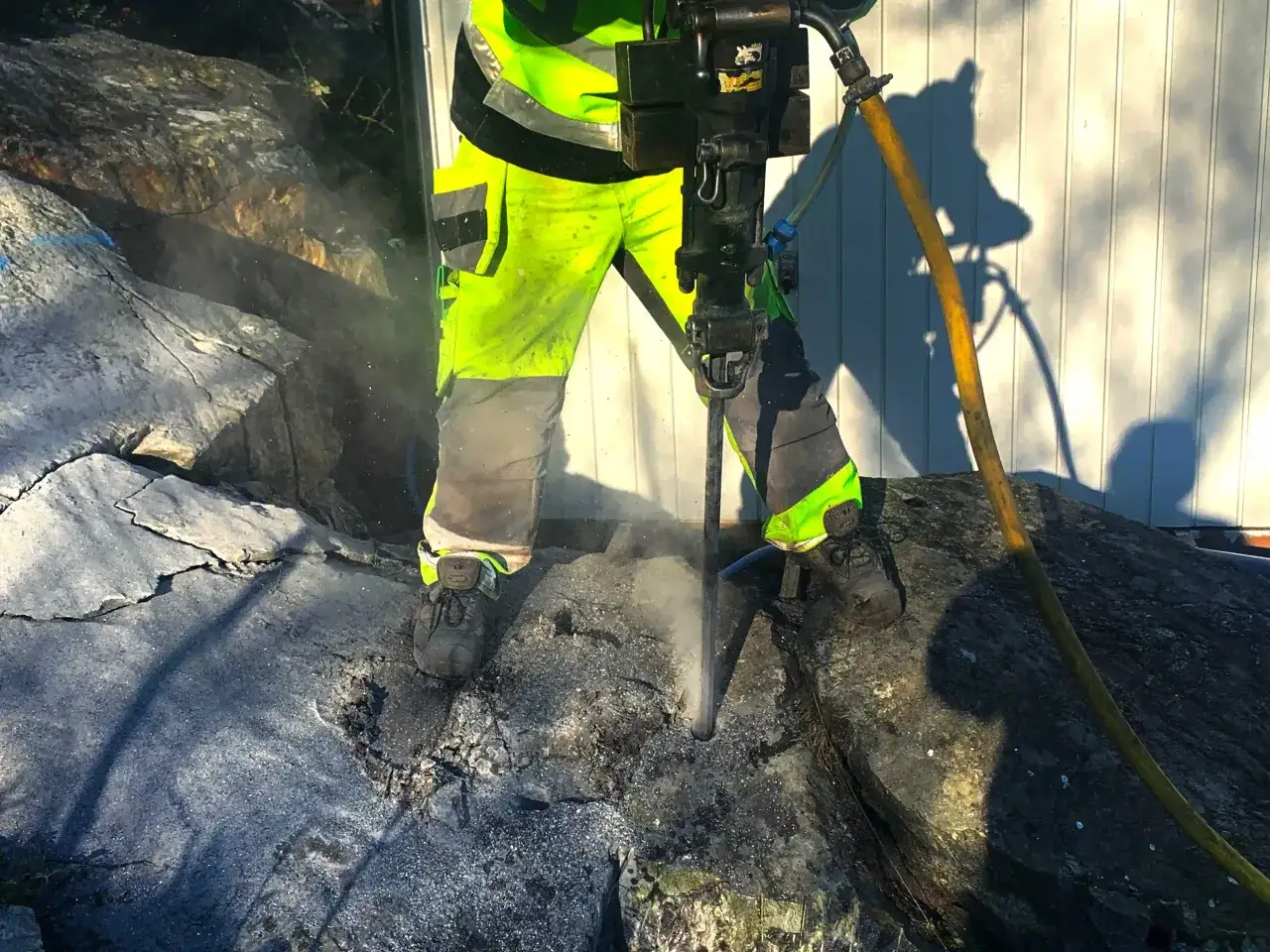 Arbetare i varselkläder borrar i berg med en kraftig borrmaskin. Dammet yr runt borren.