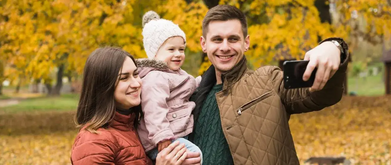 Rodzina robi selfie na tle jesiennych liści. Uśmiechnięci rodzice trzymają córeczkę, która wygląda jakby właśnie dostała babciowe.