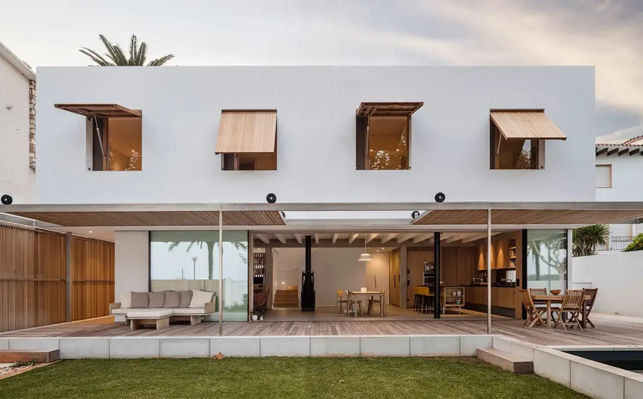 Frente fachadas de casas modernas con ventanas de madera y terraza abierta que revela un interior luminoso con cocina y comedor.
