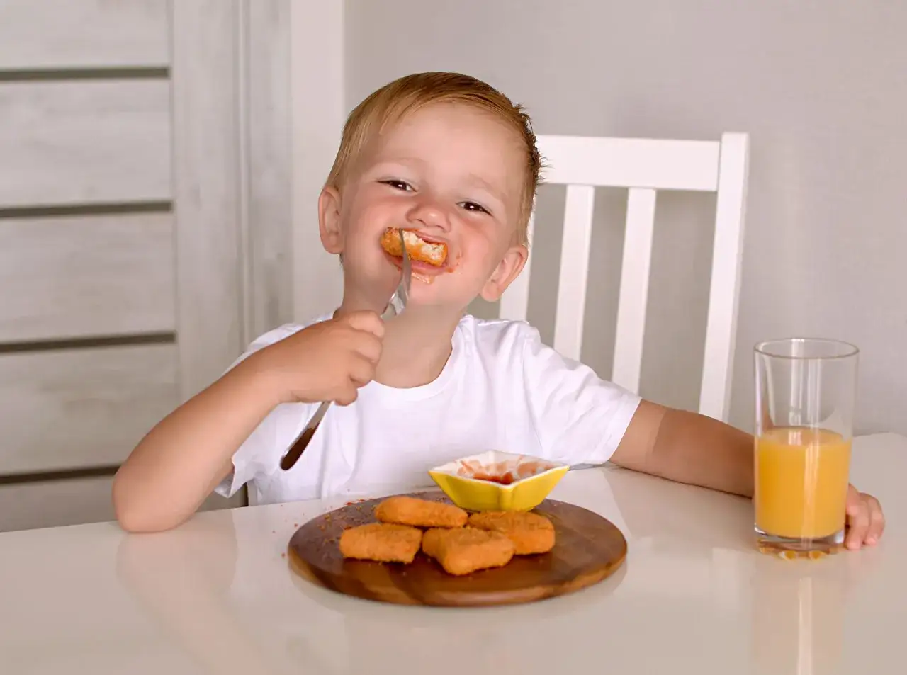 Uśmiechnięty dwulatek je nuggetsy z keczupem. To jego pyszny obiad! Obok stoi szklanka soku.