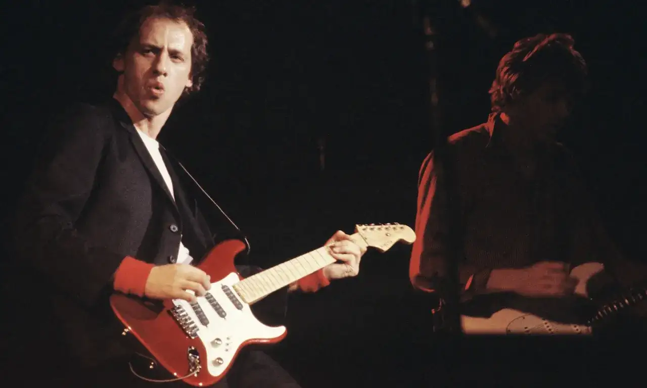 Mark Knopfler z Dire Straits gra na czerwonej gitarze, w tle drugi muzyk.