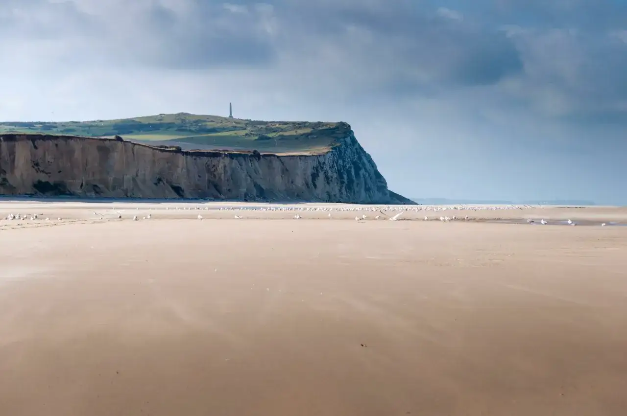 Plage de sable fin sur la C&ocirc;te d'Opale, avec des falaises imposantes et un phare au loin. Des mouettes se reposent sur le rivage.