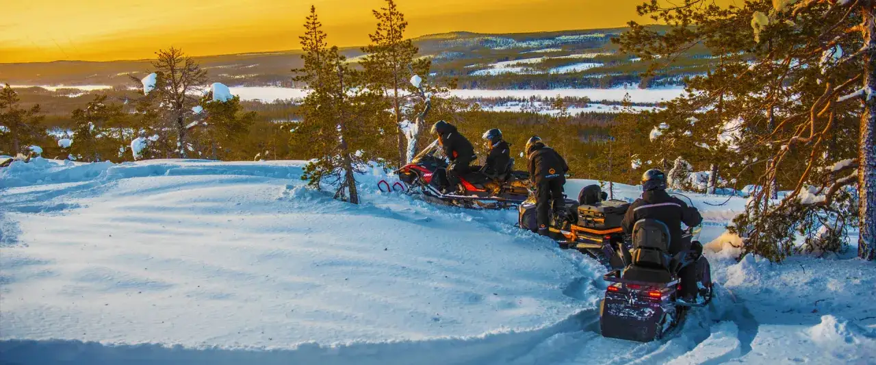 Snöskoteråkare njuter av vinterlandskapet i Norrbotten. Solnedgången färgar himlen gyllene över snötäckta träd och fjärran sjöar. Perfekta aktiviteter Norrbotten för äventyrare.