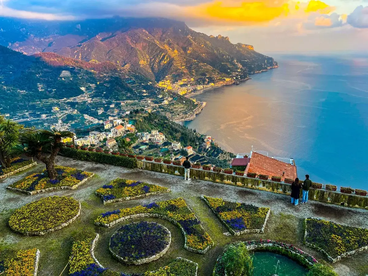 Giardini fioriti con vista mozzafiato sulla costa. Si ammira la **distanza Ravello Amalfi**, con il mare azzurro e le case aggrappate alla montagna.