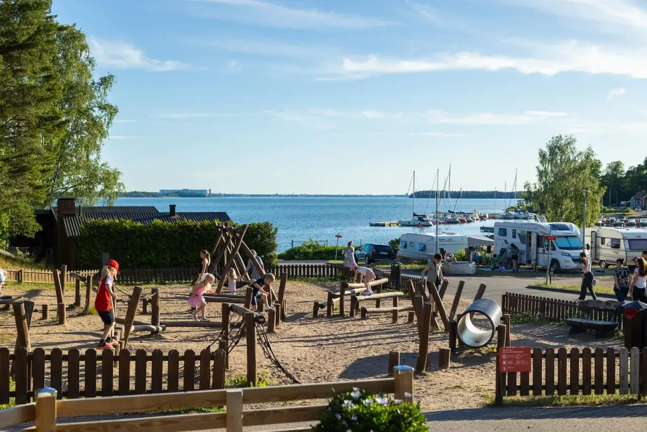 Barn leker på en sandig lekplats vid vattnet. Husbilar och segelbåtar syns i bakgrunden. Kolmården first camp erbjuder en härlig semester.