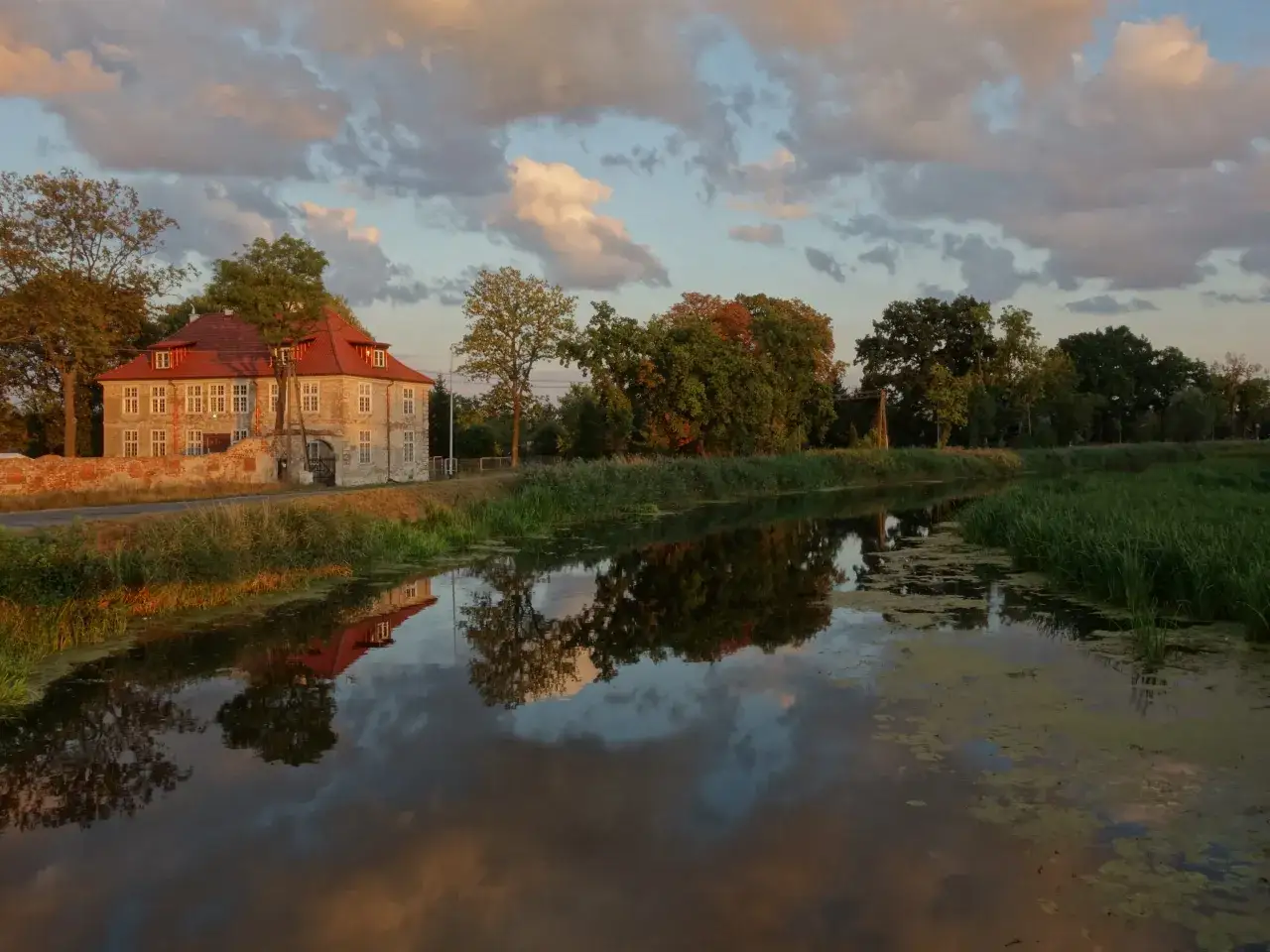 Dworek w Gdańsku Olszynce z czerwonym dachem odbija się w spokojnej wodzie kanału. Chmury na niebie malują zach&oacute;d słońca.