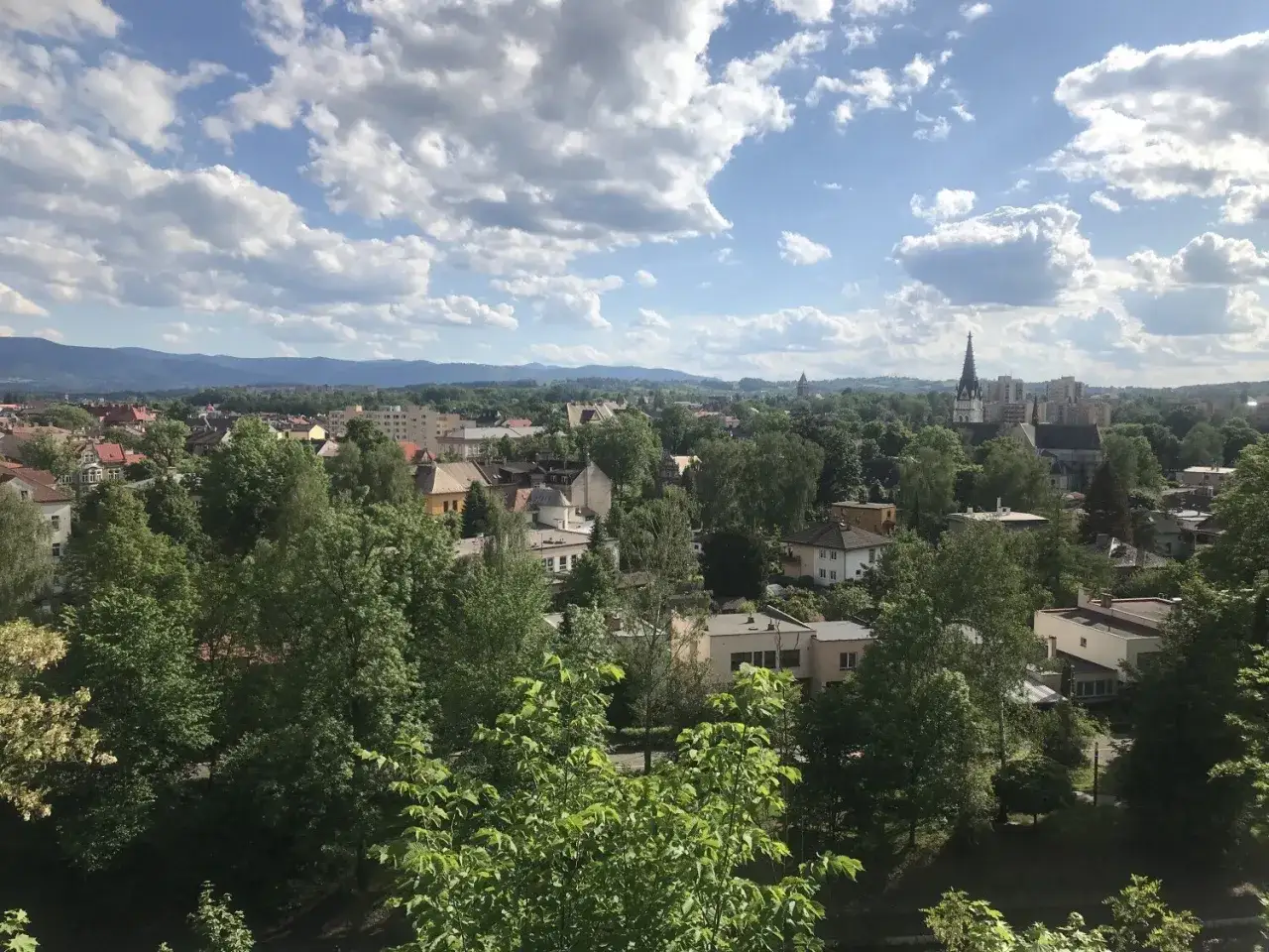 Widok na Cieszyn z góry zamkowej. Dachy domów, wieża kościelna i zielone drzewa pod błękitnym niebem z chmurami.