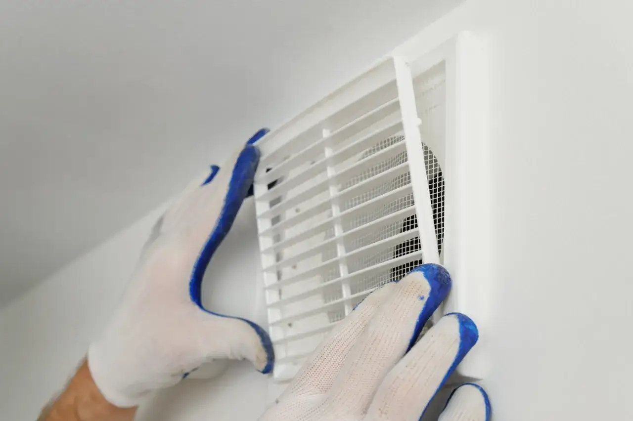 Mains gantées ajustant une grille blanche sur une bouche d'aération murale, étape clé de l'entretien VMC.